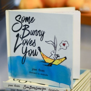 May include: A stack of children's books with a blue and white cover featuring a bunny in a banana boat. The title of the book is "Some Bunny Loves You" and the author is Jesse Thom, illustrated by Christache.