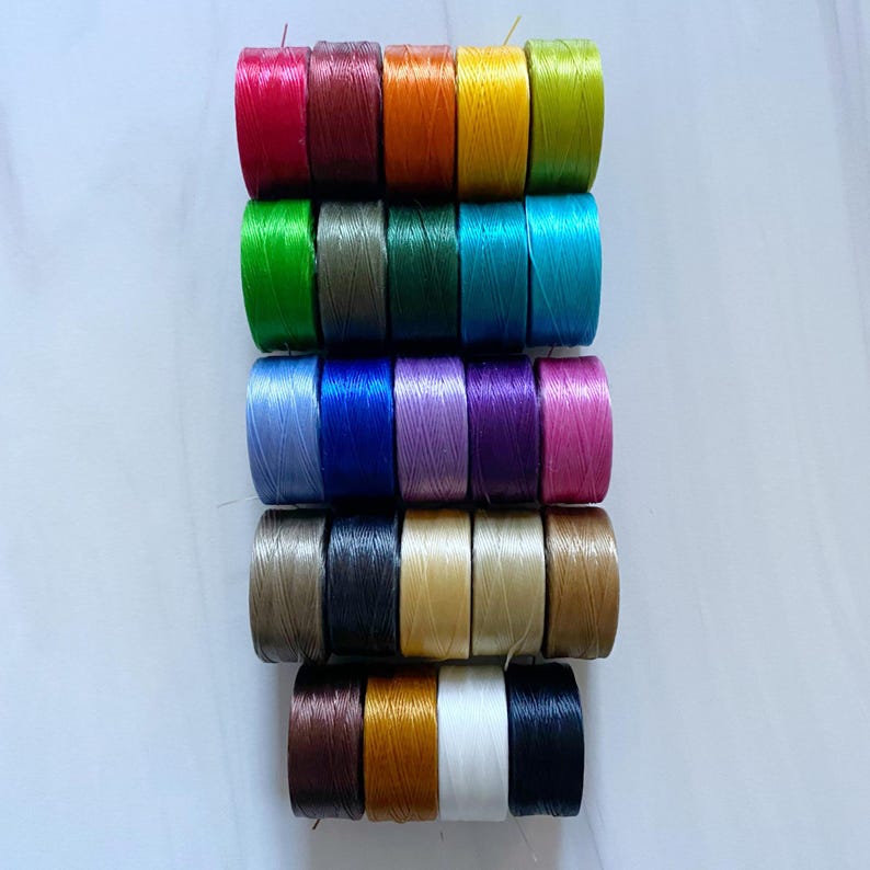 Peut inclure: Une collection de 20 bobines de fil de diff&eacute;rentes couleurs, dont le rouge, le vert, le bleu, le violet, l'orange, le jaune, le marron et le noir. Les bobines sont dispos&eacute;es en pile, la bobine du haut &eacute;tant rouge et la bobine du bas &eacute;tant noire.