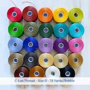 May include: Twenty-four spools of C-Lon thread in various colors, including red, orange, yellow, green, blue, purple, pink, brown, gray, and white. The spools are arranged in a grid pattern on a white surface. The text "C-Lon Thread - Size D - 78 Yards/Bobbin" is printed below the spools.