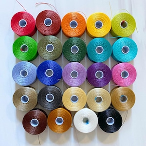 Peut inclure: Une collection de 25 bobines de fil de diff&eacute;rentes couleurs, dont le rouge, le vert, le bleu, le violet, le rose, le jaune, l'orange, le marron, le noir et le blanc. Les bobines sont dispos&eacute;es en grille sur une surface blanche.