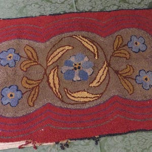 May include: A rectangular hooked rug featuring a central taupe field with a blue floral motif and yellow leaves. The rug is bordered by a wide red frame with concentric dark blue lines. The texture is thick and looped, showing a handcrafted appearance.