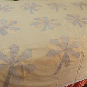 May include: A cream-coloured quilt with a repeating pattern of stylised floral designs in pale yellow and light blue. The quilt is handmade and appears to be a vintage or antique item, with a simple, elegant aesthetic.