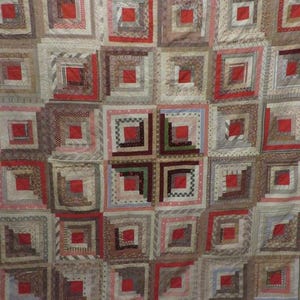 May include: A log cabin quilt featuring a repeating square block pattern. Each block contains a central solid red square surrounded by concentric strips of fabric in various neutral tones, including beige, brown, grey, and cream with diverse patterns.