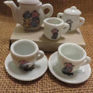 May include: A miniature white porcelain tea set with a teapot, sugar bowl, creamer, and two teacups with saucers. Each piece is decorated with a cartoon character design in various colors.