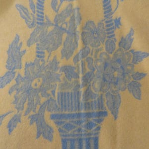 May include: A light yellow blanket with a blue floral basket design. The basket is filled with flowers and leaves. The basket has a handle and is set against a light yellow background. The design is a repeating pattern.