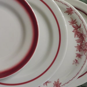 May include: Three white ceramic plates with red trim. The top plate has a solid red band around the edge. The middle plate has a thin red band around the edge. The bottom plate has a red band around the edge and a pattern of red leaves.