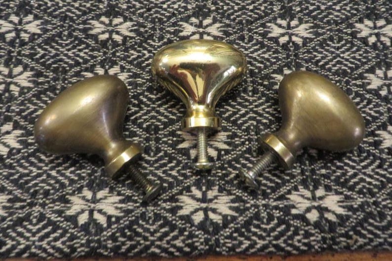 May include: Three brass cabinet knobs with a smooth, rounded, egg-shaped head and a short, cylindrical base. Each knob features a threaded metal screw extending from the bottom, designed for attachment to furniture or cabinetry doors.
