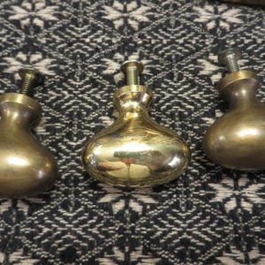 May include: Three polished brass cabinet knobs with a rounded, teardrop-shaped profile. Each knob features a smooth, reflective metallic surface and a threaded metal screw extending from the top for installation purposes.