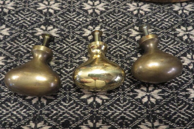 May include: Three polished brass cabinet knobs with a rounded, teardrop-shaped profile. Each knob features a smooth, reflective metallic surface and a threaded metal screw extending from the top for installation purposes.