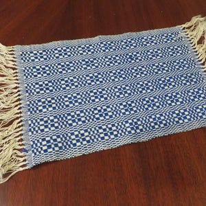 May include: A rectangular woven textile with a blue and white checkered pattern. The edges are finished with a fringe. The textile is likely a placemat or small rug.