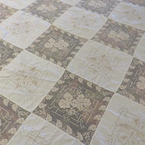 May include: A vintage quilt with a repeating pattern of alternating squares. The squares feature floral designs in cream and gray, creating a classic, textured look. The quilt is likely made of cotton or a similar fabric.