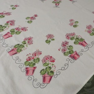 May include: A white tablecloth featuring a repeating pattern of pink potted geraniums with green leaves. The floral motifs are arranged in a border design with thin black swirling lines connecting the pots across the fabric surface.