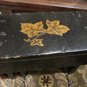 May include: A black wooden stool with a gold leaf design of a vine with leaves. The stool has a distressed finish and is sitting on a patterned rug.