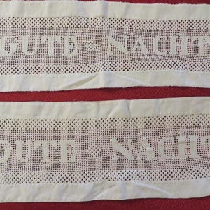 May include: Two white lace ribbons with the German words "GUTE NACHT" embroidered on them. The ribbons have a delicate, openwork design and are set against a red background.