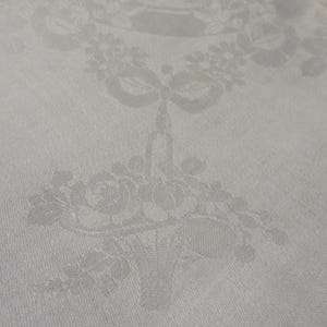 May include: A white textile fabric featuring a tonal damask weave pattern. The design displays a central basket filled with various flowers, including roses, suspended by a ribbon bow from a floral garland arrangement woven into the cloth.