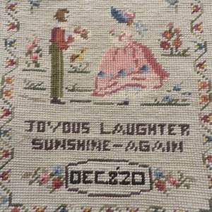 May include: A beige needlepoint with a floral border and a scene of two figures in a garden. The text "Joyous Laughter Sunshine Again" and "Dec. 20" are embroidered on the needlepoint.