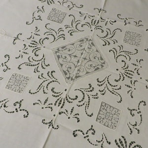 May include: White linen tablecloth with intricate cut-out designs. The design features a central square with a smaller square in each corner. The squares are filled with a delicate lace pattern.