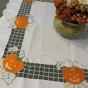 May include: A white tablecloth featuring a scalloped edge and a dark green grid-patterned border. The fabric is decorated with embroidered orange jack-o'-lantern pumpkins, green leaves, and vines. A ceramic vase filled with flowers sits on top.