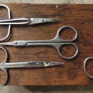 May include: Four silver-toned metal scissors with symmetrical finger loops and pointed blades. Three scissors are positioned horizontally in a stack, while one scissor is placed vertically to the right, all featuring a polished metallic finish.