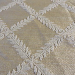 May include: A light gold fabric featuring a raised white diamond lattice pattern. Each line of the diamond grid is composed of a repeating leaf or fern motif, creating a textured, geometric design across the surface of the textile material.