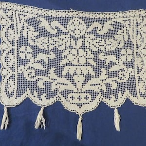 May include: A rectangular off-white lace panel featuring an intricate openwork grid pattern with a central floral motif and symmetrical leaf designs. The bottom edge has a scalloped border finished with five hanging tassels made of matching thread.