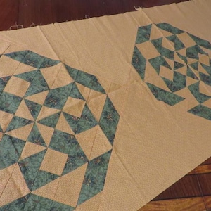 May include: A section of fabric featuring a repeating geometric pattern. The design incorporates teal and tan diamond and triangular shapes, creating a visually complex pattern. The fabric appears to be cotton or a similar material.