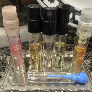 May include: A collection of perfume sample vials in a silver tray. The vials have clear glass bodies with various colored liquids and black or clear caps. Some vials have text, including "Modern Muse" and "Dot".