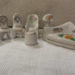 May include: A miniature porcelain dollhouse furniture set featuring a couch, four chairs, a table, and a lamp. The pieces are painted with floral designs in shades of pink, yellow, and green.