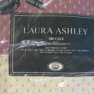 May include: A Laura Ashley twin flat sheet in a plastic package. The sheet is cream-colored with a small, repeating floral pattern in burgundy. The packaging includes the brand name, product details, and dimensions.