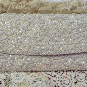 May include: A white beaded clutch purse with a floral design. The purse is made of small, iridescent beads and has a flap closure.