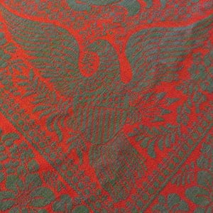 May include: Close-up of a red textile with a repeating pattern of a stylized bird and floral elements in a contrasting green color. The fabric has a textured appearance, suggesting a woven or embroidered design.