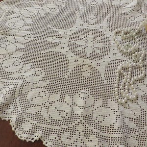 May include: White crocheted doily with a floral design. The doily is round and has a scalloped edge. A strand of white pearls is lying on top of the doily.