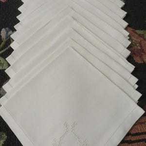 May include: A stack of off-white linen napkins, each with a decorative embroidered design in the corner. The napkins are neatly arranged, showcasing their square shape and delicate stitching. The background is a dark floral pattern.