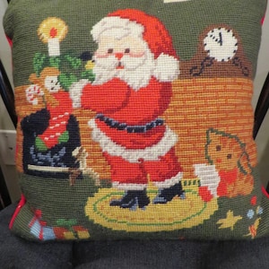 May include: Needlepoint Christmas pillow featuring Santa Claus in a red suit, white beard, and hat. The pillow has a green background with a fireplace, clock, and a dog. The pillow is trimmed with red piping.