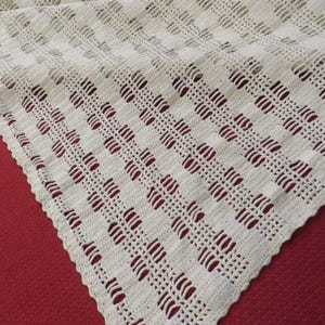 May include: Cream-colored, crocheted blanket with a repeating openwork pattern. The blanket is triangular in shape and has a scalloped edge. The texture of the crochet work is visible, and the blanket is displayed on a red surface.