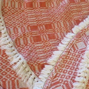 May include: Close-up of a woven textile with a geometric pattern in shades of orange and white. The edges are trimmed with a white fringe, adding a textural contrast to the design. The fabric's intricate weave suggests a handcrafted item.