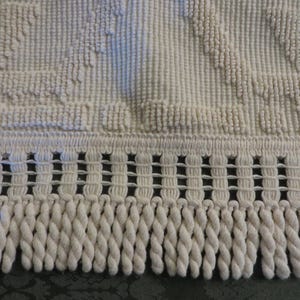 May include: Close-up of a cream-colored, textured rug with a woven border and knotted fringe. The rug has a pattern of raised, textured shapes. The fringe is made of thick, twisted cords.