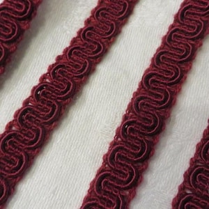 May include: Close-up of four burgundy-colored decorative trim ribbons with a looped design. The ribbons are arranged diagonally against a white, textured background. The trim is likely used for sewing or crafting projects.