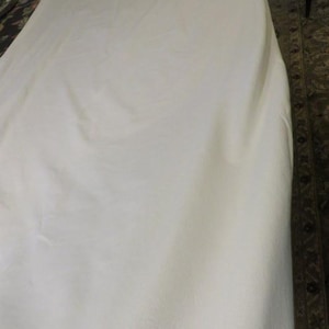 May include: A large, off-white sheet of fabric, possibly linen or cotton, is draped over an unseen object. The fabric has a soft, slightly textured appearance with subtle folds and shadows. The background includes a patterned rug and a portion of a dark chair.