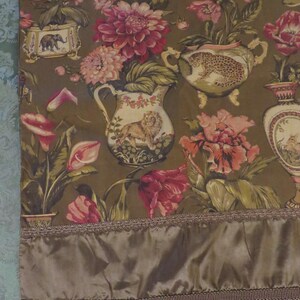 May include: A decorative fabric panel with a repeating pattern of floral arrangements and animal illustrations in vases. The colour palette includes shades of brown, pink, and green. The bottom edge has a satin border with fringe.