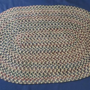 May include: An oval-shaped braided rug featuring a multi-colored pattern of interwoven fabric strips in shades of tan, dark blue, burgundy, and cream. The rug is constructed with a continuous concentric braided design that creates a textured surface.