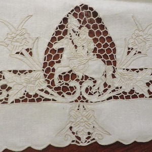 May include: Cream-colored embroidered textile with a central design of a person on horseback, surrounded by floral and geometric patterns. The embroidery is detailed, with a scalloped edge along the bottom.