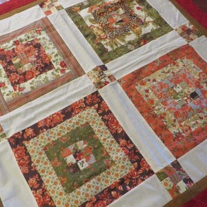 May include: A handmade quilt with a patchwork design. The quilt features four large square blocks with floral patterns in shades of red, orange, green, and brown, separated by white fabric. The quilt is trimmed with a brown border and a red edge.