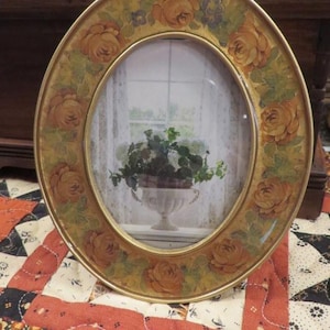 May include: An oval picture frame with a gold-toned metal border featuring a painted floral pattern of orange roses and green leaves. The frame contains a print of a potted green plant in a white urn set against a window with curtains.