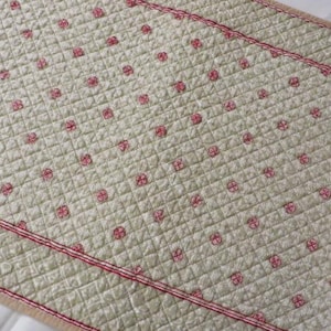 May include: Cream-colored quilted table runner with a repeating pattern of small red floral designs. The edges are trimmed with a red stitched border. The runner is made of a soft, textured fabric.