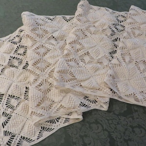 May include: Off-white, crocheted shawl with a geometric diamond pattern. The shawl has a fringed edge and is folded over, showing the intricate openwork design. The texture is soft and the color is a creamy white.