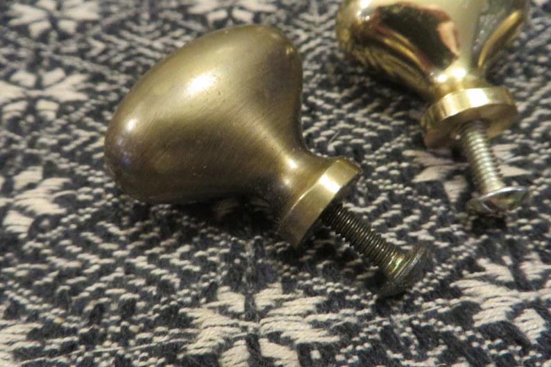May include: Two brass cabinet knobs with a smooth, rounded, oval-shaped head and a cylindrical base. Each knob features a threaded metal bolt extending from the back, designed for attachment to furniture or cabinetry doors and drawers.