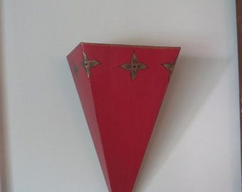 May include: A wall-mounted sconce featuring a triangular, inverted pyramid shape in a solid red finish. The top edge is decorated with a repeating pattern of four-petaled floral motifs in a metallic gold color, arranged horizontally across the front.
