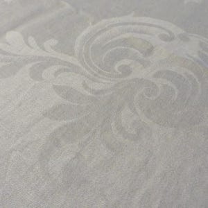 May include: Close-up of a white fabric with a subtle, embossed floral pattern. The design features swirling, stylized leaves and scrollwork, creating a textured surface. The fabric appears to be a light, possibly linen or cotton, material.