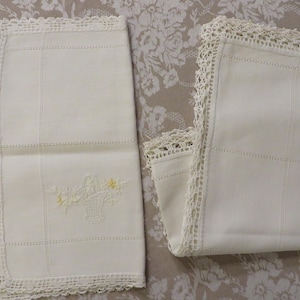 May include: Two off-white linen napkins with crocheted lace edges. One napkin features an embroidered basket of flowers with yellow accents. The napkins have a grid pattern of stitched lines.